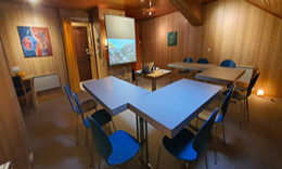 La salle aménagée pour une conférence : tables en U, chaises bleues et écran de projection